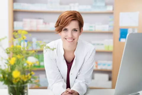 Eine Drogistin lehnt am Tresen in der Apotheke und lacht in die Kamera